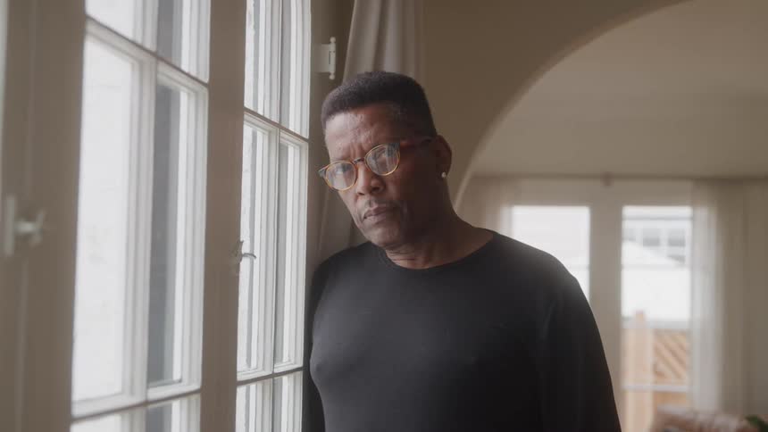 The man leans against the windows in his room