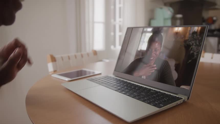 A person is engaged in a video call on a silver laptop which is placed on a wooden table on the screen a woman with dark hair and a grey top is seen actively participating in the conversation