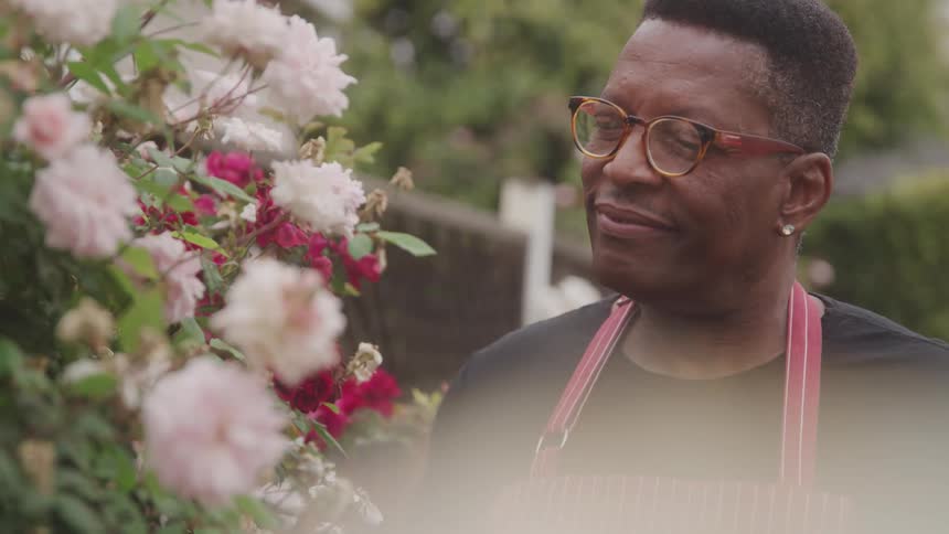 A man with short dark hair and glasses wearing a black shirt and a red and white striped apron is shown smiling gently he is standing outdoors next to a blooming rose bush with light pink and red flowers