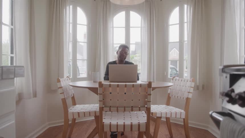 Man sitting at his dining table near a splay bay window during the day uses a laptop