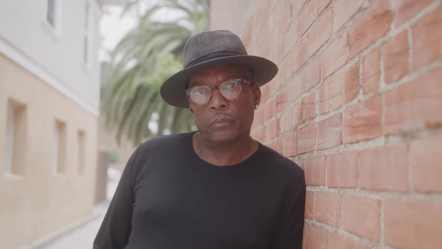 A man wearing a black longsleeved shirt a grey fedora and glasses is leaning against a brick wall he is looking directly at the camera with a neutral expression