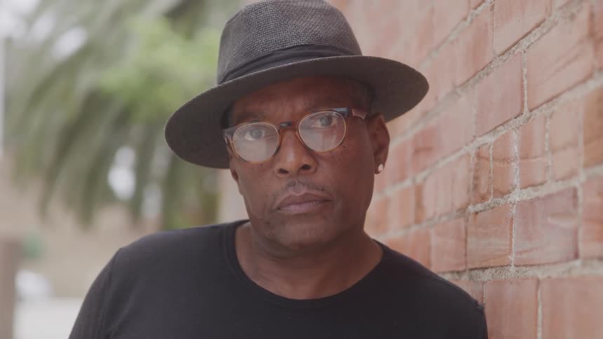 A man wearing a grey fedora hat and glasses is leaning against a brick wall he has a serious expression and is looking directly at the camera