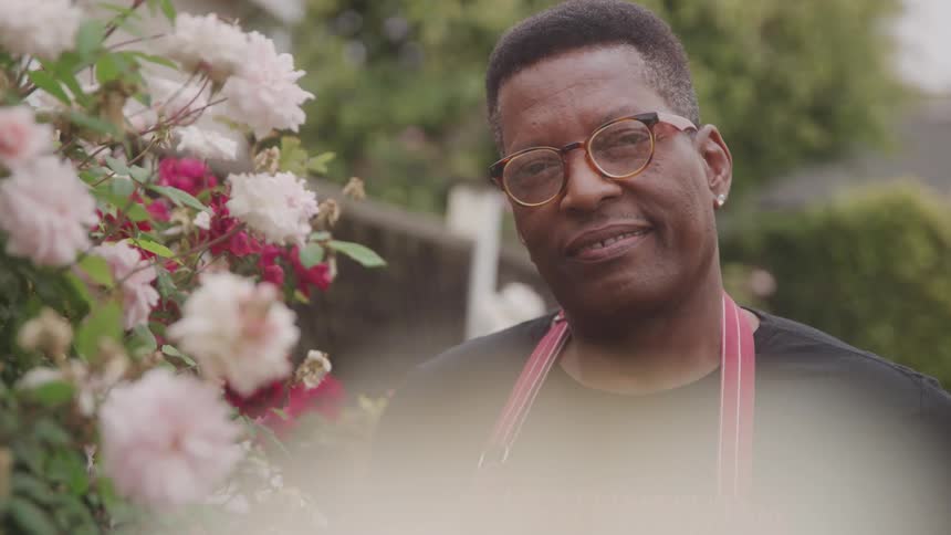 A middleaged man stands in a garden at midday beside a potted flower plant wearing glasses an apron and earrings with a smile on his face