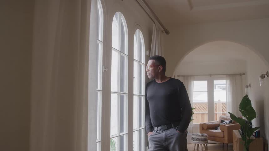 A man in a black longsleeved shirt and grey pants stands by a large window with multiple arched panes he is looking out thoughtfully with his hand in his pocket while the living room with a couch is visible in the background