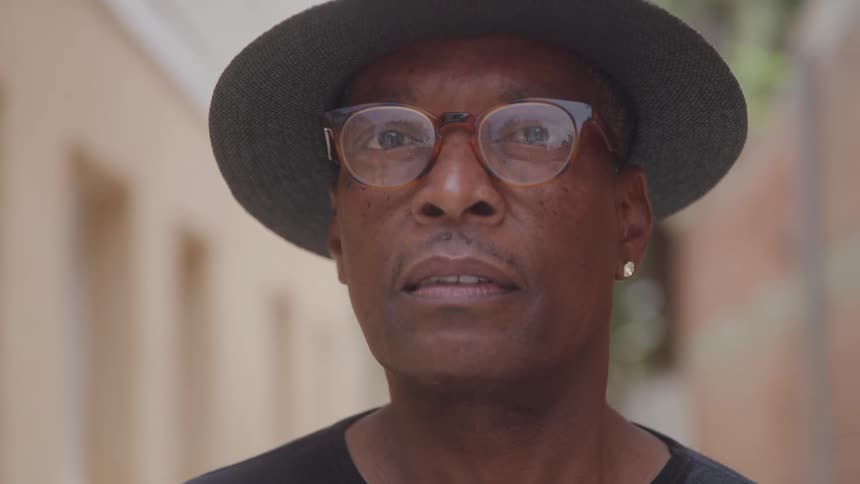 Man in a hat and glasses turns his head slowly to face the camera