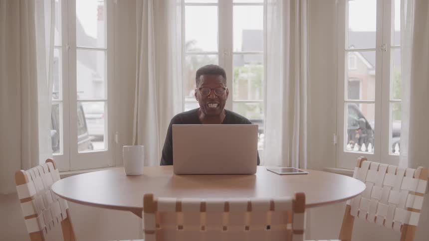 A man wearing glasses and a dark shirt is seated at a round wooden table working on a laptop he smiles and waves at the camera while a white mug and a tablet rest on the table beside his laptop with large windows providing natural light in the backgrou