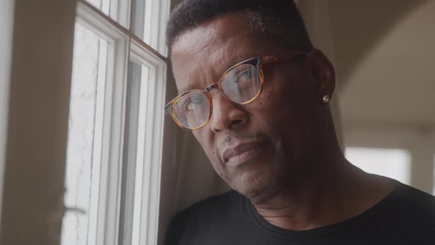 A middleaged black man wearing an earring glasses and a black tshirt rests his head beside a window indoors at midday