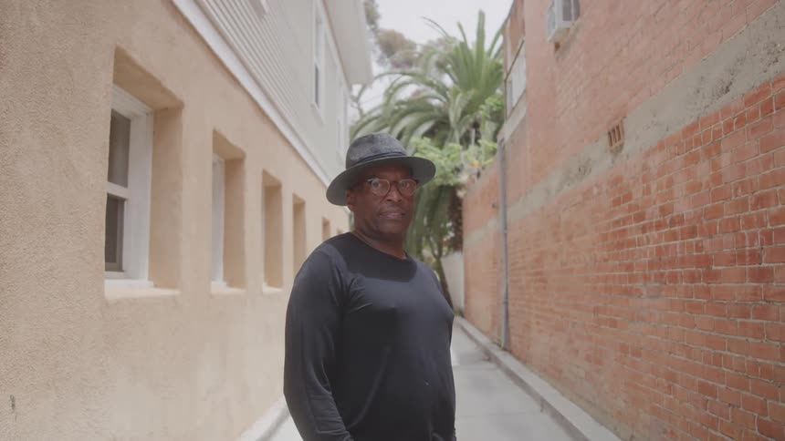 A man stands behind an apartment building at midday wearing a hat glasses and a black shirt