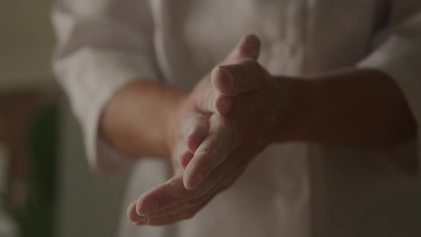 A closeup shot shows a persons hands covered in white powder likely flour as they rub them together small particles of the powder are visibly falling from their hands as they rub