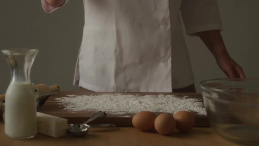 A chef pours flour onto a surface in preparation for making dough with three eggs a glass of milk and a bowl on the side