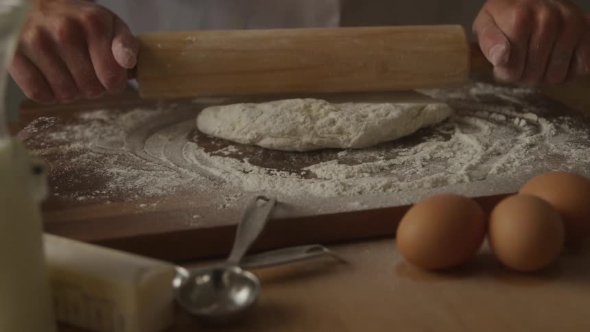 A persons hands are seen using a wooden rolling pin to flatten a dough on a floured wooden surface nearby baking ingredients such as three eggs a stick of butter measuring spoons and a bottle of milk are visible suggesting a baking activity in prog
