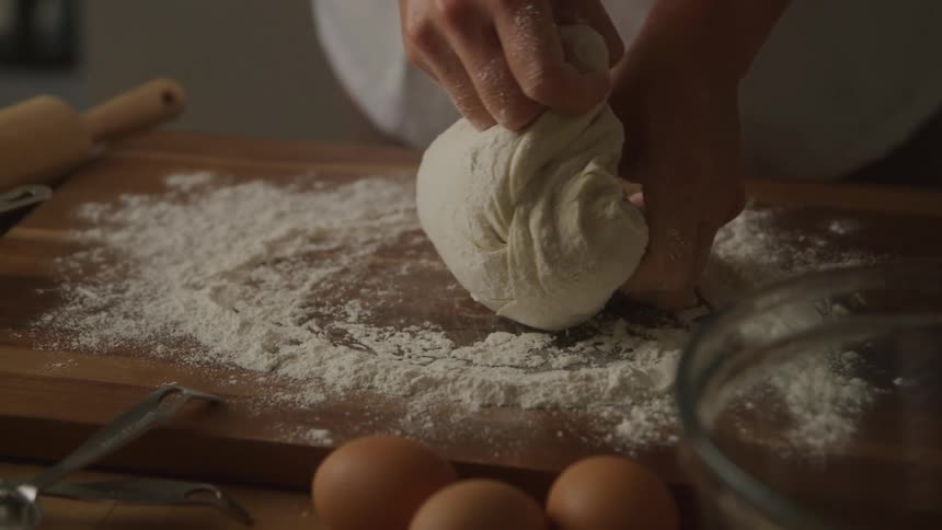 A person molds dough on a wooden surface with three eggs on the side