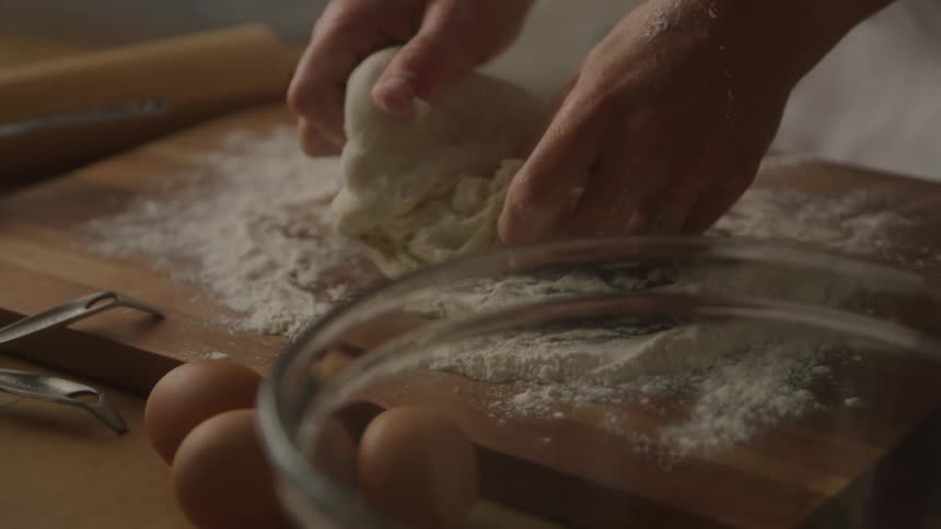 A person molds dough with bare hands on a wooden surface with three eggs and a glass bowl in front and a rolling pin on the left side