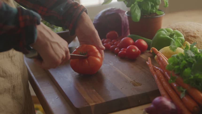A closeup of hands precisely slicing a fresh red tomato on a wooden cutting board amidst an array of colorful vegetables