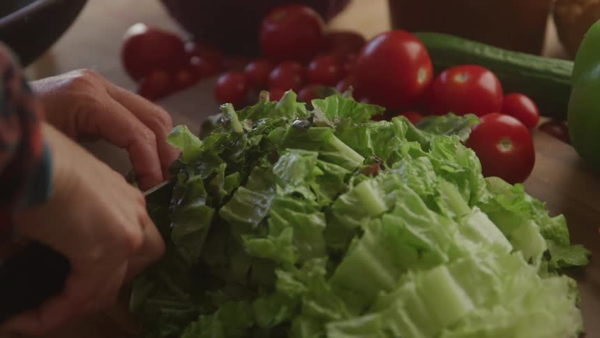 A persons hands chop green lettuce on a wooden cutting board with fresh red tomatoes and green vegetables blurred in the background