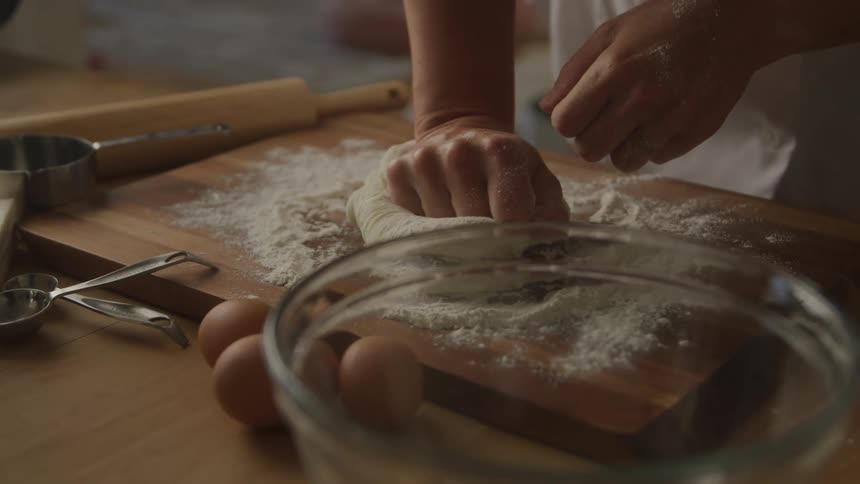 A person is kneeding a dough by using his hands and some flour and there are some eggs and a bowl