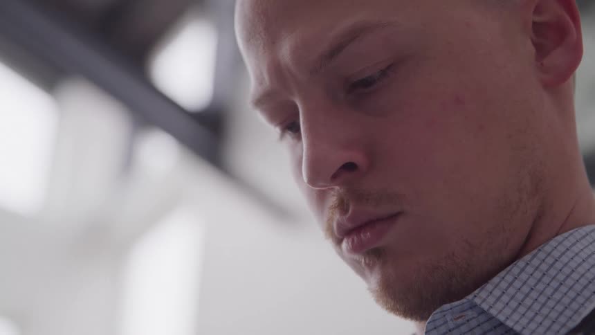 A closeup shot of a man with a shaved head and a checked shirt looking downwards with a pensive expression