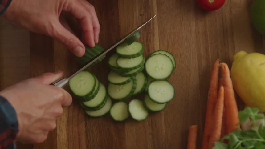 A topdown view of hands expertly slicing a green cucumber into thin rounds on a wooden cutting board surrounded by other fresh produce