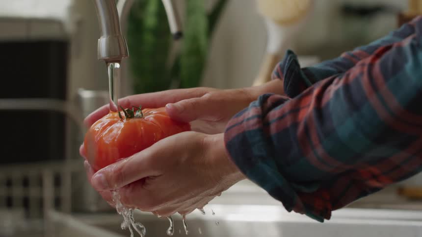 A closeup of hands meticulously rinsing a large textured tomato under a steady stream of water in a kitchen sink