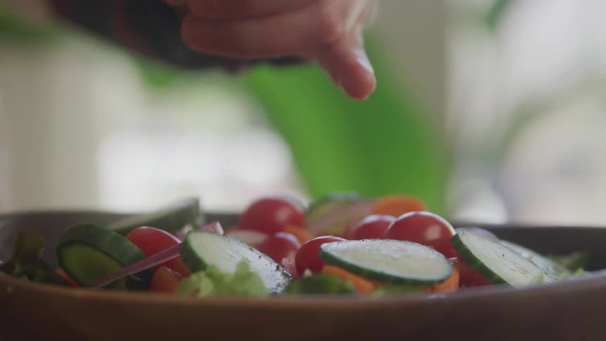 A person slices various vegetables into a bowl and adds spices on top