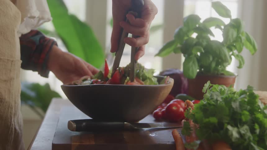 A closeup of hands actively tossing a vibrant fresh salad in a dark bowl with tongs on a rustic wooden surface surrounded by other wholesome ingredients