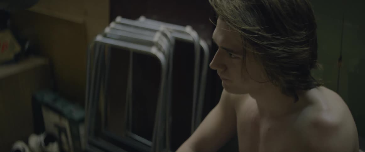A bare chested man sits and frowns