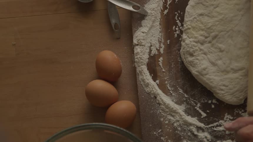 A flat lay shot displays a wooden cutting board dusted with flour beside which rest three brown eggs and a glass bowl a rolling pin lies parallel to a mound of dough suggesting preparations for baking