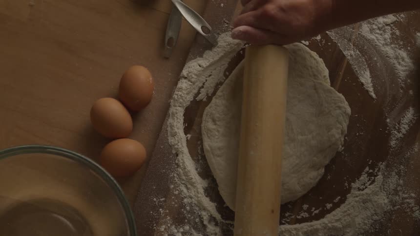A person is seen from a topdown perspective using a wooden rolling pin to flatten a lightcolored dough on a wooden surface dusted with flour three brown eggs and a clear mixing bowl are visible beside the dough suggesting a baking activity