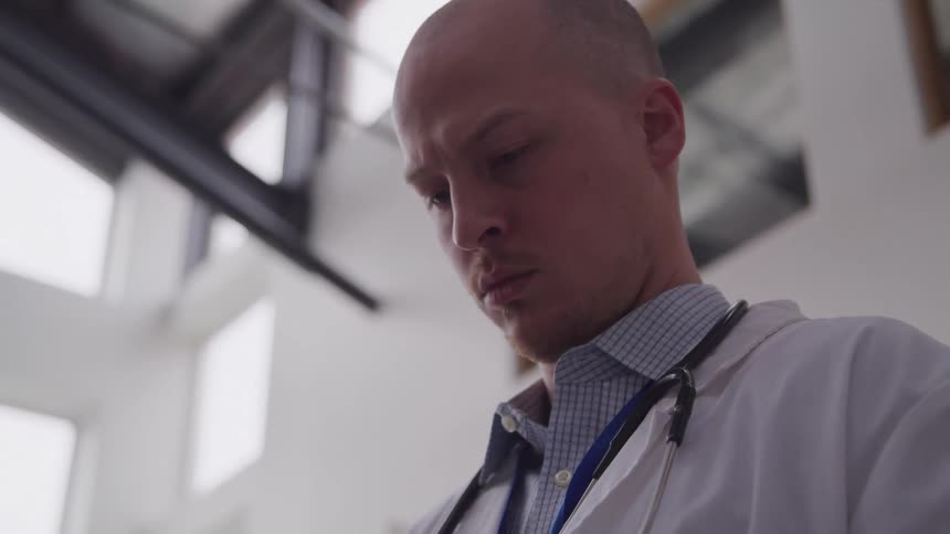 A serious bald doctor in a white lab coat with a stethoscope around his neck looks intently downwards engrossed in a task