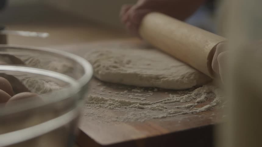A person is seen using a wooden rolling pin to flatten a dough on a floured wooden surface in the background a clear bowl and a container of milk are visible suggesting a baking activity is underway