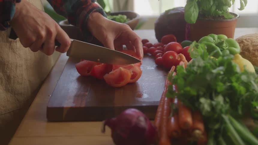 A person slicing a tomato on a cutting board with a knife as other vegetables surround the table at midday