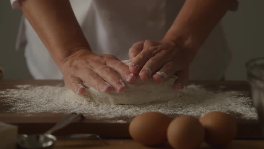A chef seen from the waist up wearing a white uniform kneads a mound of dough on a wooden board sprinkled with flour with three brown eggs nearby