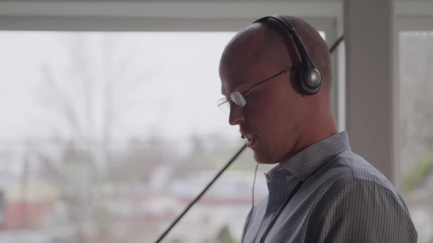 A young man wearing a headset and glasses walks indoors at midday operating his phone just beside a glass window