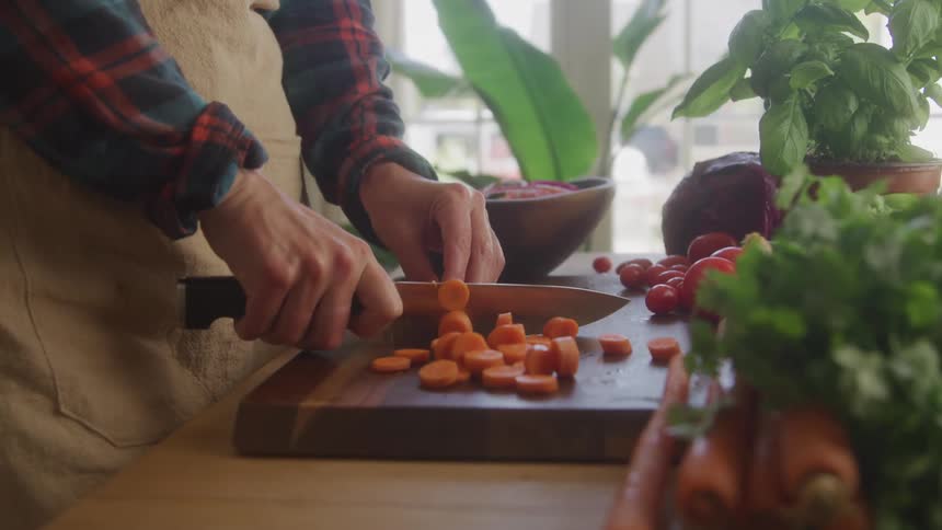 A lowangle closeup of hands rhythmically chopping bright orange carrots on a wooden cutting board surrounded by other fresh produce in a sunlit kitchen