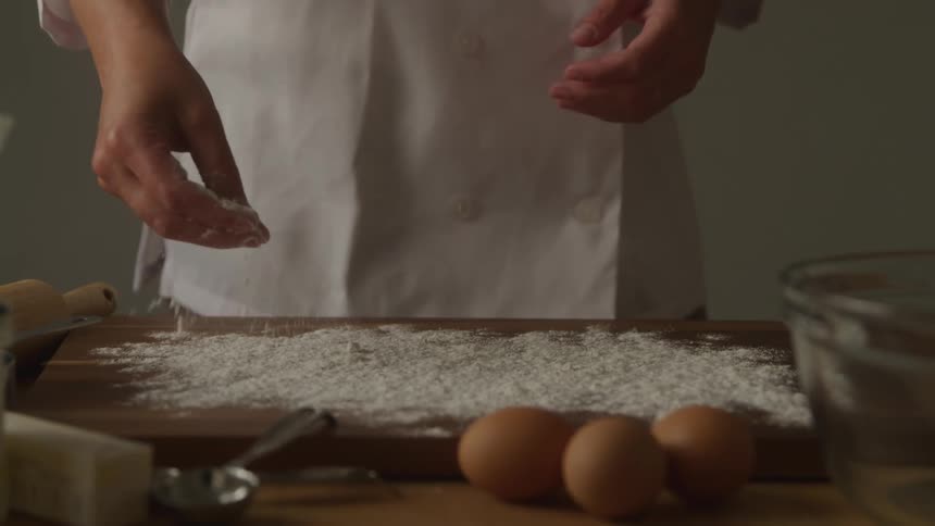A chef pours flour onto a rolling board with three eggs and a bowl in front