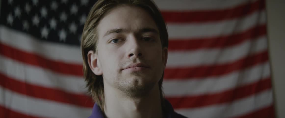 Man stoically looking into the camera with an american flag draped behind him
