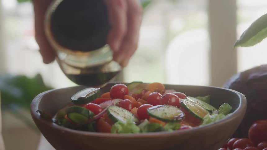 A hand pours sauce from a container into a bowl filled with a variety of sliced vegetables