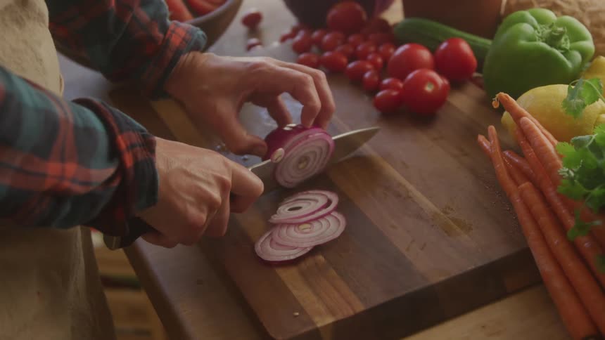 A person wearing a plaid shirt is carefully slicing a red onion on a wooden cutting board with thinly sliced pieces already visible various fresh vegetables including tomatoes bell peppers carrots and lemons are artfully arranged in the background