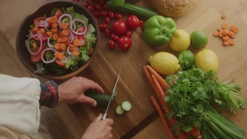 A person cuts vegetables and places them in a bowl filled with a variety of vegetables while other uncut vegetables remain on the table waiting to be cut