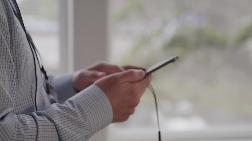 A person in a lightcolored longsleeved shirt with a lanyard around their neck is holding a darkcolored tablet or large smartphone looking at its screen they appear to be wearing wired earphones with the wires dangling from the device