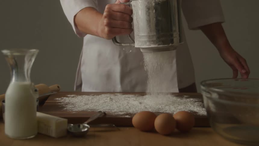 A persons hands are shown sifting flour onto a wooden board surrounded by baking ingredients like eggs milk and butter