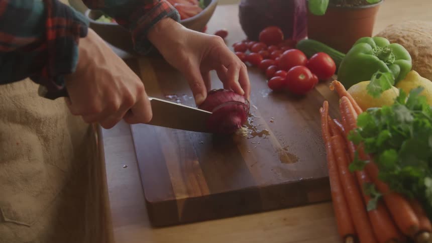 A pair of hands is shown carefully chopping a red onion on a wooden cutting board surrounded by fresh vegetables the scene is brightly lit and focuses on the detailed action of preparing a healthy meal