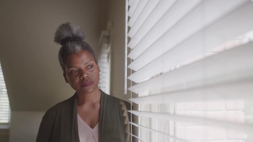 An older adult woman with her grey hair in a bun wearing a green cardigan and a light colored top looks out a window through white blinds with a somber expression