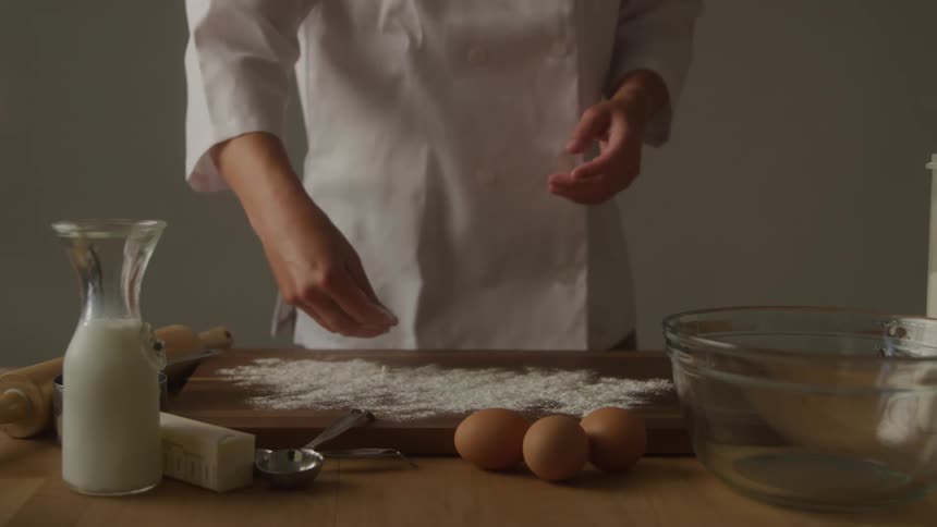 A chef in a white uniform stands at a wooden counter sprinkling flour onto a cutting board with ingredients such as milk butter and eggs neatly arranged around her