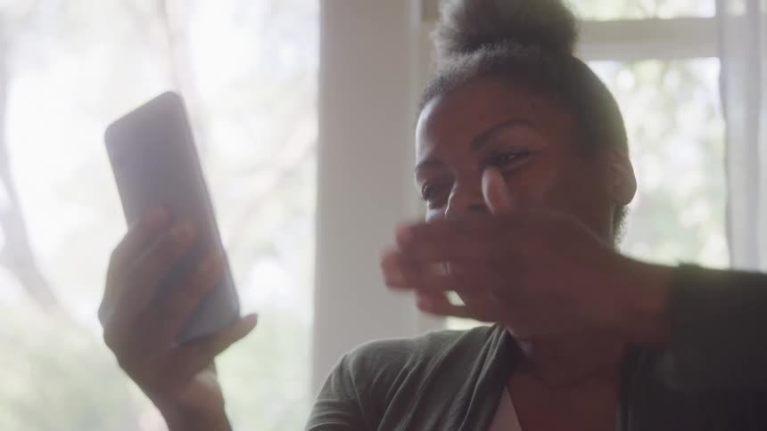 A young woman holding her phone and waving while smiling during a video call indoors at midday
