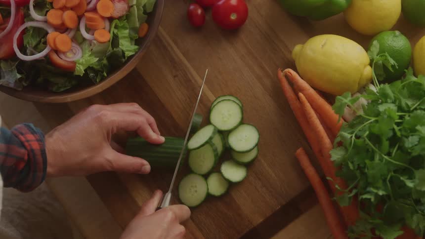 A topdown view of hands expertly slicing a green cucumber into thin rounds on a wooden cutting board surrounded by other fresh produce