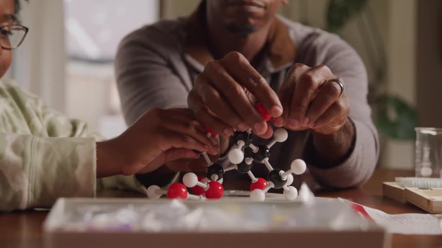 A black man dismantles an organic chemistry model with his daughter on a table indoors during daytime