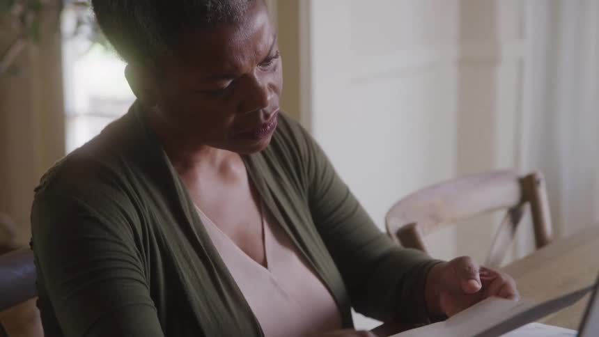 An older woman with short grey hair wearing a green cardigan and a light colored top sits at a table and reads a document with a concerned expression with a laptop and computer mouse in front of her