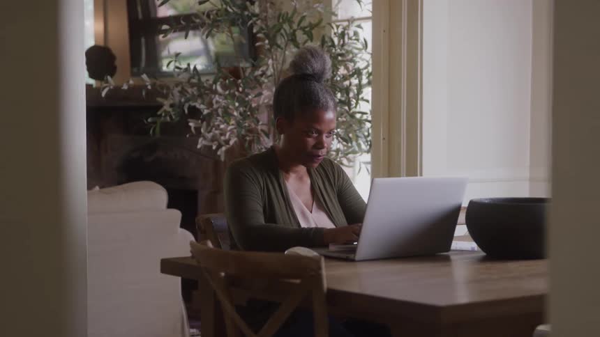 A woman sits at the kitchen table in her home typing on a laptop