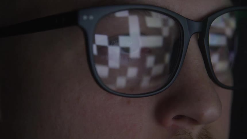 A person wearing blackframed glasses is shown in a closeup with digital pixelated reflections on the lenses suggesting they are looking at a screen with changing images or data the foreground shows their eyes behind the glasses and the background i